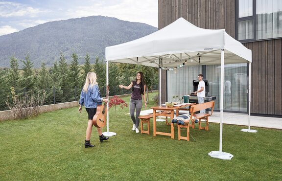 Der Faltpavillon steht im Garten und ist für die Gartenparty bereit. Die Frau kommt mit einer Gitarre und wird von der Gastgeberin empfangen. Unter dem Gartenzelt steht ein Tisch für die Gartenparty.