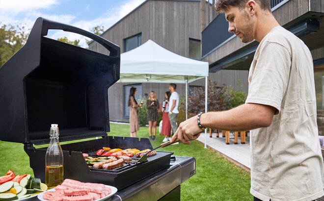 Man is grilling meat and vegetables in the garden. Behind him the guests are standing under the grey folding pavilion. They are drinking a glass of wine.