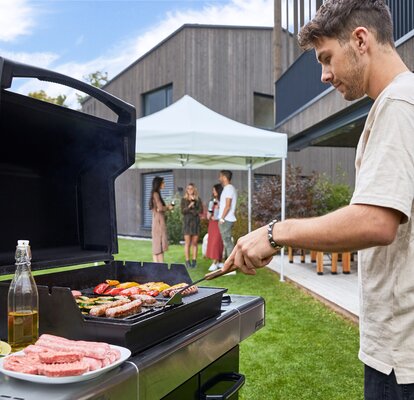 Mann grillt im Garten Fleisch und Gemüse. Dahinter stehen die Gäste unter dem grauen Faltpavillon und trinken ein Glas Wein.