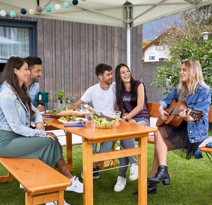 Freunde sitzen unter dem Faltpavillon auf einer Rustica-Garnitur und lachen. Ein Mädchen spielt mit der Gitarre.