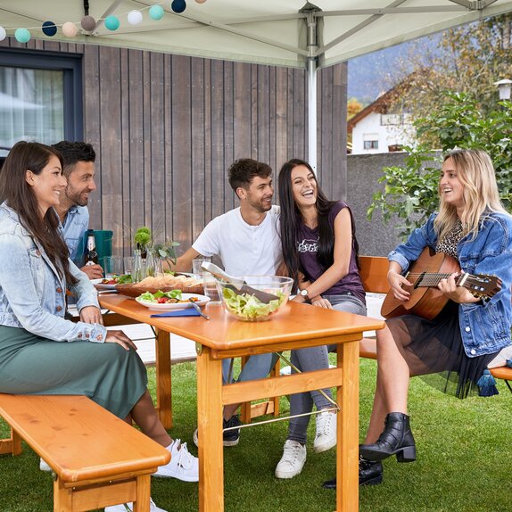 Freunde sitzen unter dem Faltpavillon auf einer Rustica-Garnitur und lachen. Ein Mädchen spielt mit der Gitarre.