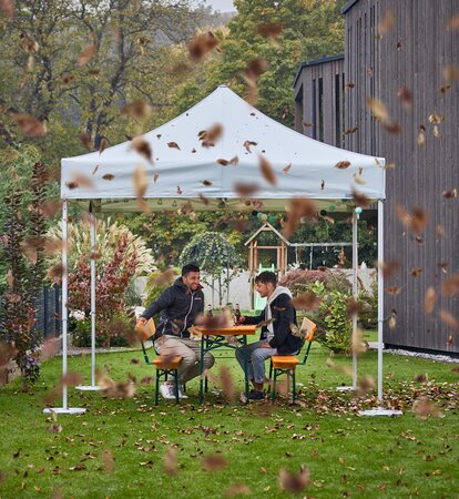 Zwei Männer sitzen bei starkem Wind im Garten unter einem Faltpavillon. Überall wirbeln Blätter.