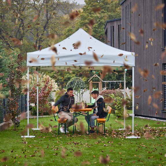 Due uomini sono seduti sotto un gazebo pieghevole con del vento forte che soggia. Ci sono dell foglie che volano. 