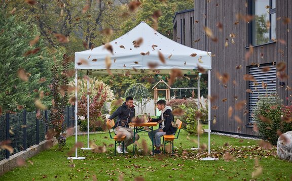 Two men are sitting in the garden under a gazebo. Leaves are swirling everywhere.