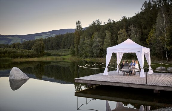 Weißer Faltpavillon steht auf dem Steg am See. Darunter dinieren 4 Personen.