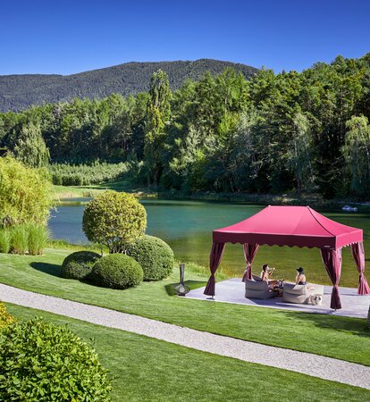 Ein eleganter bordeauxroter Faltpavillon steht direkt am See. Darunter sitzt ein Paar auf zwei Liegestühlen und genießt einen Drink.