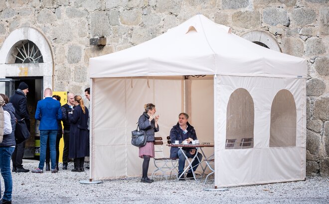 The smoking tent with 3 side walls is located in front of the restaurant. Below it a woman who smokes and a man who drinks a coffee are talking. 
