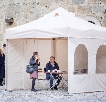 The smoking tent with 3 side walls is located in front of the restaurant. Below it a woman who smokes and a man who drinks a coffee are talking. 