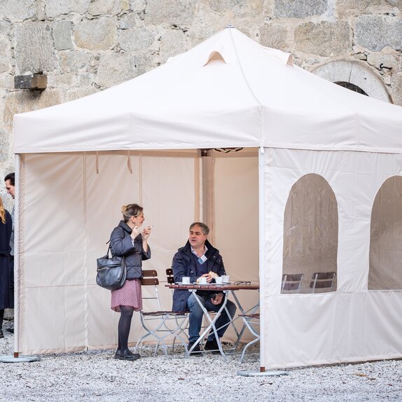 Il gazebo pieghevole per fumatori di fronte al ristorante ha 3 pareti laterali. Sutto stanno conversando una donna che fuma e un uomo che beve un caffè.