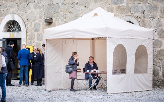 Il gazebo pieghevole per fumatori di fronte al ristorante ha 3 pareti laterali. Sutto stanno conversando una donna che fuma e un uomo che beve un caffè.