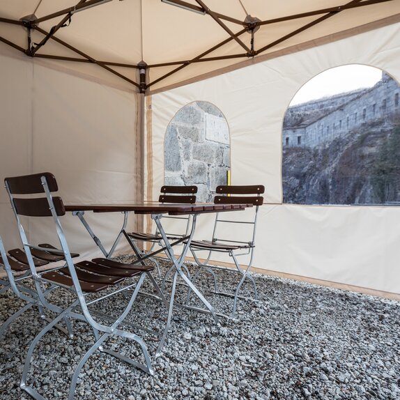 Der Faltpavillon von innen. Es steht ein Tisch mit Stühlen im Zelt. Man kann die Seitenwand mit Fenstern sehen.