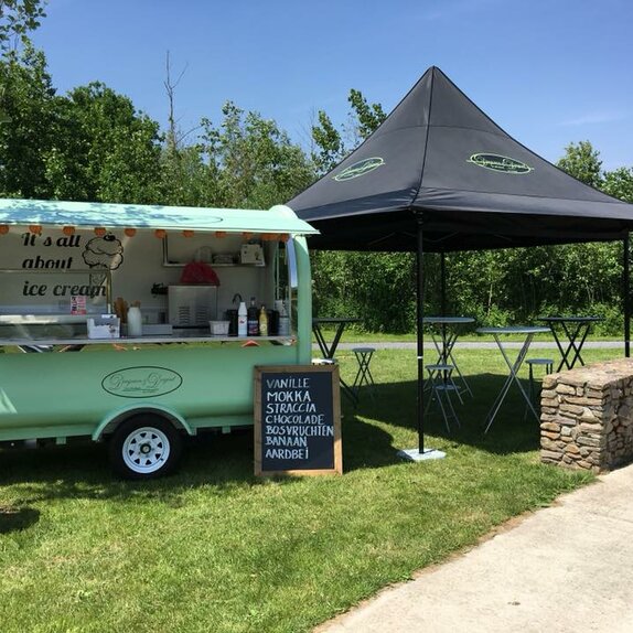 Der Street Food Stand von "Dingenen & Dergent" steht auf einer Wiese. Der türkisfarbene Anhänger steht neben dem schwarzen Faltpavillon.