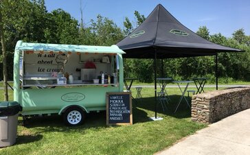 The street food stand of "Dingenen & Dergent" is located on a meadow. The turquoise trailer is standing next to the black folding pavilion.