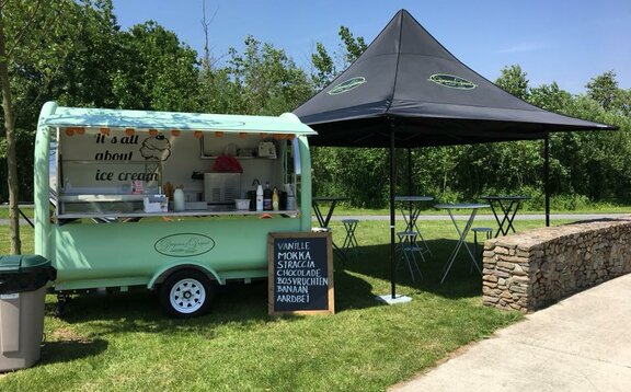 Der Street Food Stand von "Dingenen & Dergent" steht auf einer Wiese. Der türkisfarbene Anhänger steht neben dem schwarzen Faltpavillon.