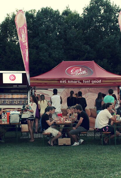 The bordeaux-red folding pavilion of "Eat the Ball" is placed next to the food truck. People are sitting in front of it. They are eating their burgers.
