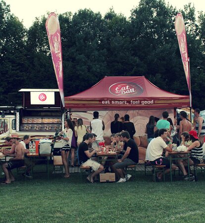 The bordeaux-red folding pavilion of "Eat the Ball" is placed next to the food truck. People are sitting in front of it. They are eating their burgers.