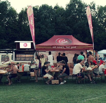 Il gazebo pieghevole color bordeaux di "Eat the ball" si trova di fianco al furgone. Di fronte ci sono delle persone che mangiano un hamburger.