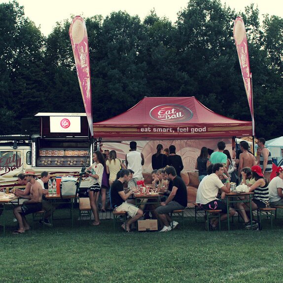 Il gazebo pieghevole color bordeaux di "Eat the ball" si trova di fianco al furgone. Di fronte ci sono delle persone che mangiano un hamburger.