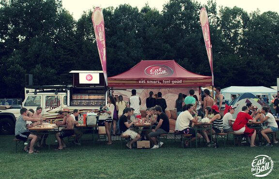 The bordeaux-red folding pavilion of "Eat the Ball" is placed next to the food truck. People are sitting in front of it. They are eating their burgers.