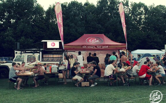 The bordeaux-red folding pavilion of "Eat the Ball" is placed next to the food truck. People are sitting in front of it. They are eating their burgers.