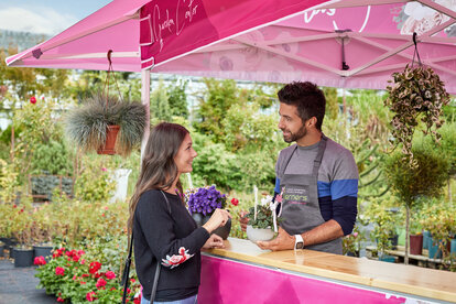 Verkaufsgespräch zwischen dem Verkäufer der Gärtnerei und einer Kundin unter einem pinken Faltpavillon mit Vordach.