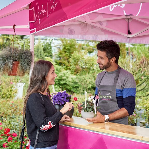 Verkaufsgespräch zwischen dem Verkäufer der Gärtnerei und einer Kundin unter einem pinken Faltpavillon mit Vordach.