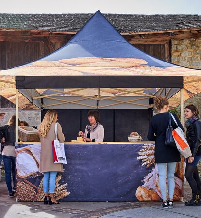 Das Marktzelt ist für eine Bäckerei und ist daher mit verschiedenem Brot bedruckt. Dahinter steht die Verkäuferin und schneidet ein Brot zur Verkostung an.