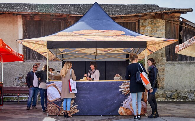 A piaci sátor egy pékség számára készült, ezért különböző kenyérfajtákkal van nyomtatva. Mögötte az eladónő egy kenyeret szeletel kóstoláshoz.