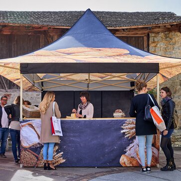 The market tent is for a bakery and is therefore printed with different types of bread. Behind it is standing the saleswoman cutting a loaf of bread for tasting.