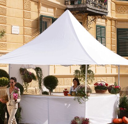 Der Marktstand für Blumen ist weiß und hat vier Vordächer auf allen Seiten. Die Kundin entfernt sich gerade vom Stand und hält eine Blume in der Hand.