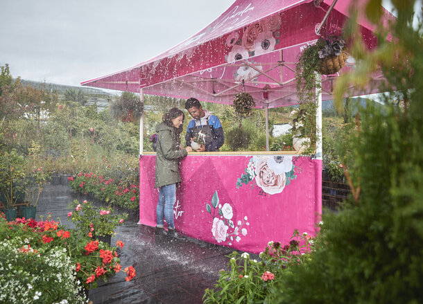 In pink und rosa bedruckter Faltpavillon mit Vordach auf einem Markt. Der Verkäufer zeigt der Kundin die Blume unter dem Vordach. Es regnet in Strömen.
