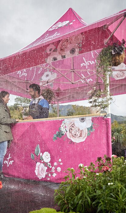 Bedruckter Faltpavillon mit Vordach im Regen. Die Kundin kauft beim Blumenhändler eine Blume. Es regnet und sie steht unter dem Vordach.