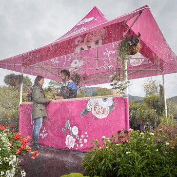 Bedruckter Faltpavillon mit Vordach im Regen. Die Kundin kauft beim Blumenhändler eine Blume. Es regnet und sie steht unter dem Vordach.