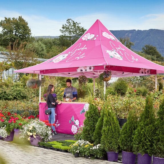 Der Faltpavillon mit Vordach ist vollflächig bedruckt. Darunter verkauft der Verkäufer gerade Blumen an eine Frau.