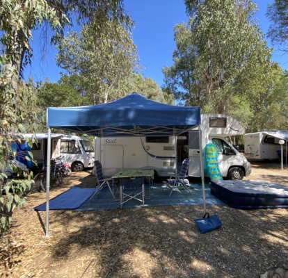 Blue camping tent 3x3 m stands in front of the caravan on the campsite. Under it there is a table with chairs.