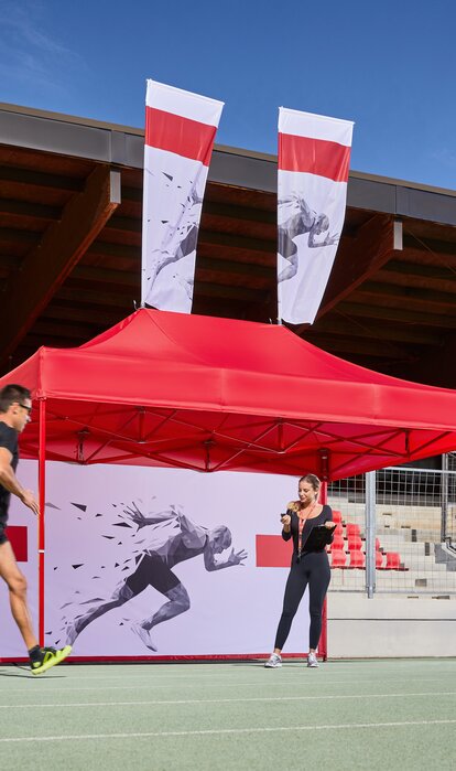 Un gazebo rosso per club sportivi e eventi sportivi si trova su una pista da corsa. Un corridore sta correndo sulla pista. Gazebo con stampa personalizzata Mastertent 4,5x3m con parete laterale. 