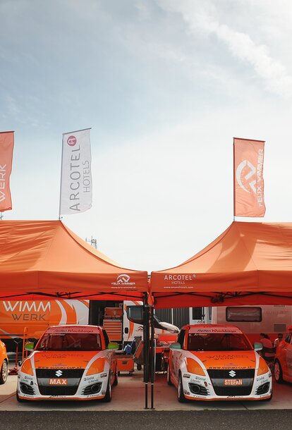 Zwei orange Paddockzelte vom Team Wimmer Werk stehen nebeneinander. Darunter stehen die Autos des Rennstalls.