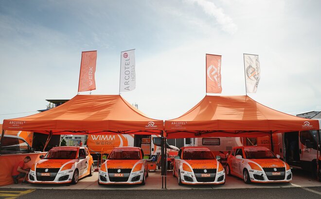 Două corturi portocalii din padocul echipei Wimmer Werk se află unul lângă celălalt. Dedesubt se află mașinile echipei de curse.