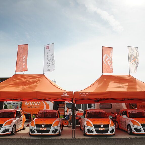 Zwei orange Paddockzelte vom Team Wimmer Werk stehen nebeneinander. Darunter stehen die Autos des Rennstalls.