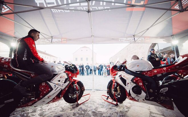 Two red motorcycle racers are standing with their bikes under the gazebo. 