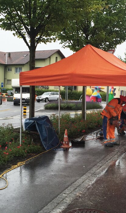 An orangegazebo is standing at a construction site. Below it are two construction workers who are carrying out drilling work.