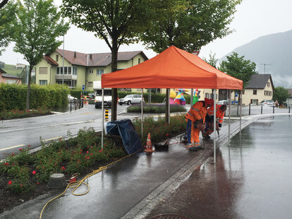 Un gazebo pieghevole arancione su un cantiere. Sotto il gazebo pieghevole ci sono due operai che lavorano.