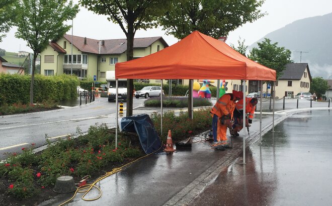 An orangegazebo is standing at a construction site. Below it are two construction workers who are carrying out drilling work.