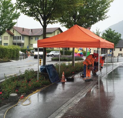 An orangegazebo is standing at a construction site. Below it are two construction workers who are carrying out drilling work.