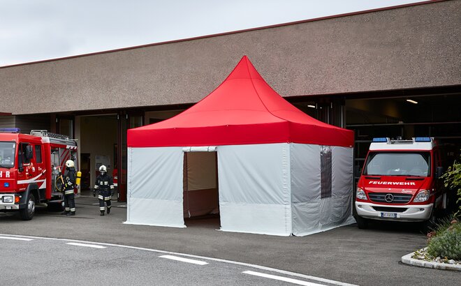 A firebrigade gazebo with red roof and grey side walls is standing in front of the building. Next to it are 2 fire engines. The firefighters are just coming out of the building. 
