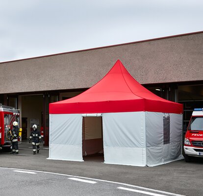 A firebrigade gazebo with red roof and grey side walls is standing in front of the building. Next to it are 2 fire engines. The firefighters are just coming out of the building. 