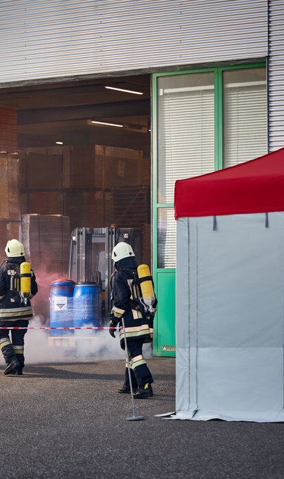 The firemen are carrying a victim of a chemical accident out of the hall. They are bringing the injured person to the gazebo. 