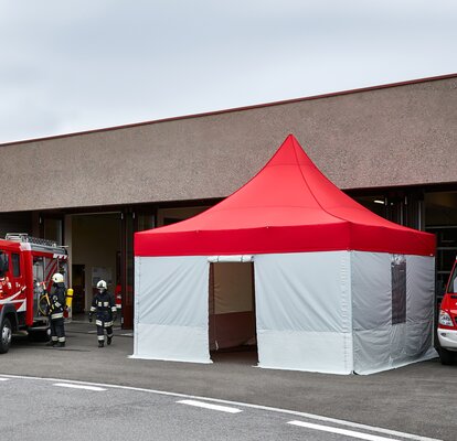 Das rote Pagodenzelt steht vor dem Gebäude und dient der Feuerwehr als Einsatzzelt. Neben dem Pagodenzelt steht ein Feuerwehrauto.