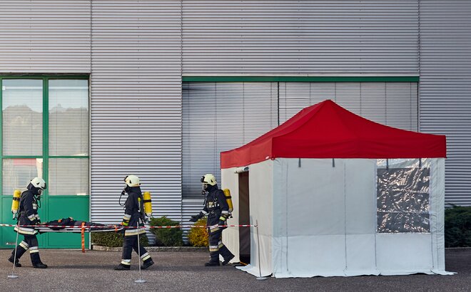 The firemen are carrying the injured person to the rescue tent. The rescue tent has a red roof and grey side walls. 