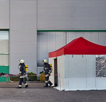 Die Feuerwehrmänner tragen das Unfallopfer in das Rescue-Zelt. Das Rescue-Zelt hat ein rotes Dach und graue Seitenwände.